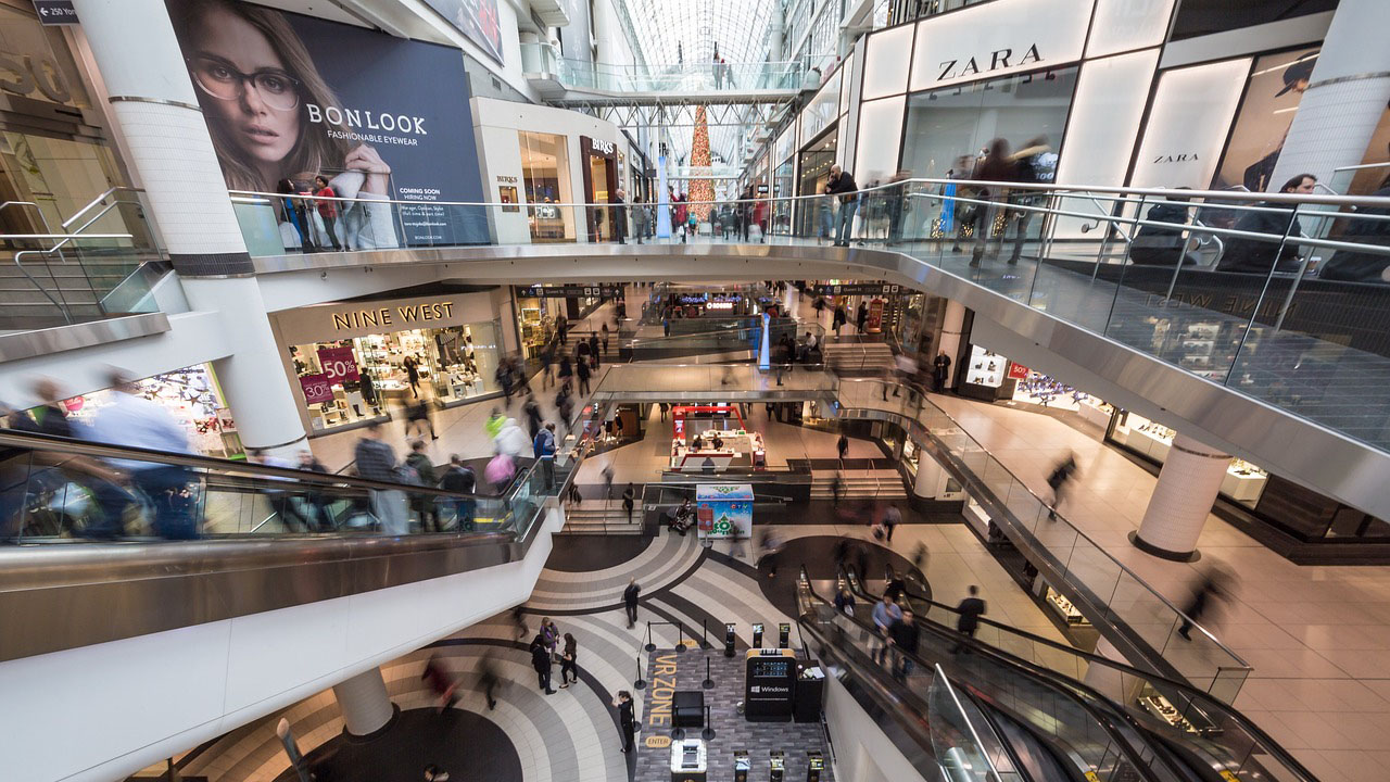 A large, busy mall with meany people walking and on escalators, featuring stores like Nine West and Zara